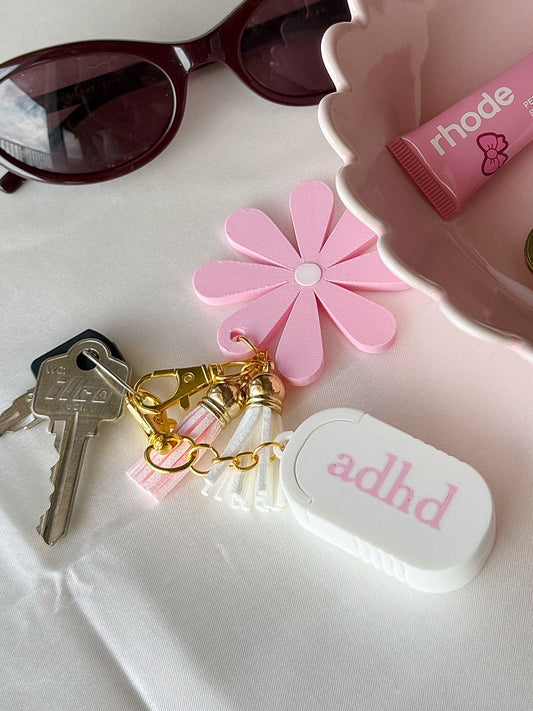 Pink lip balm, sunglasses, keys, and a small container with 'adhd' on a white surface.